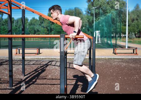 Ein junger Mann wird an einem sonnigen Tag in einem Outdoor-Workout-Bereich Dips an parallelen Bars gezeigt. Er konzentriert sich auf seine Übung und demonstriert Stärke Stockfoto