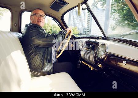 Ein älterer Mann in einer schwarzen Jacke auf einer Stadtstraße, der einen Oldtimer fährt Stockfoto