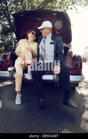 Ein älterer Ehemann und eine ältere Frau sitzen im Kofferraum eines alten Autos und halten Hände Stockfoto