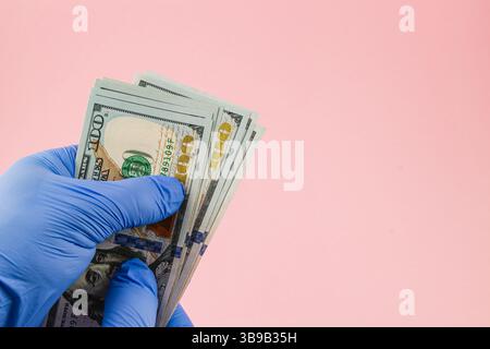 Eine Hand mit einem blauen Handschuh hält mehrere hundert Dollar Scheine vor einem sanften rosa Hintergrund, was die Hygiene und Sicherheit beim Umgang mit Bargeld unterstreicht. Stockfoto