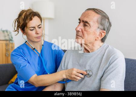 Junge Ärztin, die zu Hause einem älteren Patienten Herzgeräusche zuhört Stockfoto