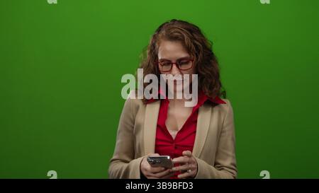 Frau mit Smartphone vor isoliertem grünen Hintergrund, die rote Bluse und einen beigefarbenen Blazer mit roter Brille in einem Studio-Setting trägt, in dem Technik dargestellt wird Stockfoto