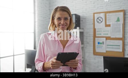 Frau lächelt drinnen mit Tablet am Arbeitsplatz mit heller Beleuchtung und moderner Einrichtung. Stockfoto
