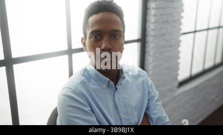 Der junge Mann in einem Büro mit Tageslicht durch große Fenster strahlt Selbstvertrauen und Professionalität aus, der in einem modernen Arbeitsbereich mit zwangloser Sitzgelegenheit sitzt Stockfoto
