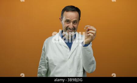 Mann in weißem Fell, der Zahnaligner über orangem Hintergrund hält, Vertrauen und Expertise ausstrahlt, reifer Profi, der modernste Orthodo zeigt Stockfoto