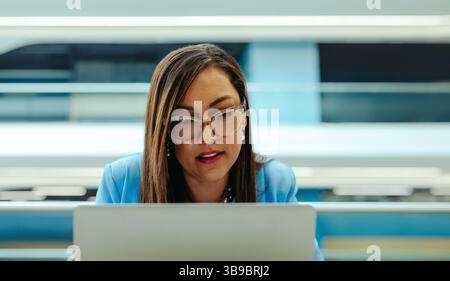 Eine fokussierte Geschäftsfrau in Brille und blauem Blazer arbeitet an ihrem Laptop in einer modernen Büroumgebung. Das Bild fängt ihre Konzentration und Professi ein Stockfoto