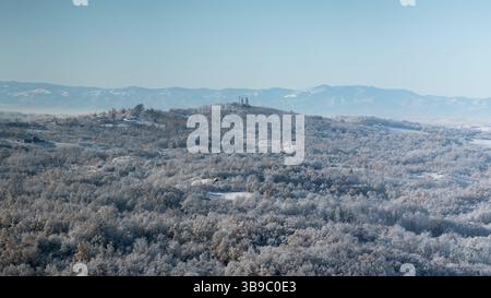 Hügeliges Dorf bedeckt mit Frost nach der kalten Winternacht, es gibt Kirche auf dem Hügel umgeben von Laubwald, Bergkette in der Ferne Stockfoto