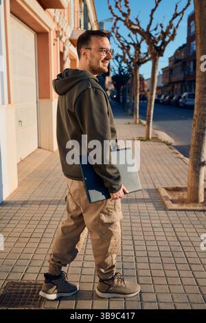 Lächelnder Mann, der auf dem Bürgersteig mit Laptop im Wohnbereich läuft Stockfoto
