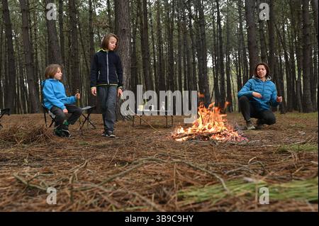 Mutter und zwei Kinder sitzen am Lagerfeuer im Wald, essen und entspannen an einem kleinen Tisch. Sie genießen ein gemütliches Essen im Freien und die Umgebung für die ganze Familie Stockfoto