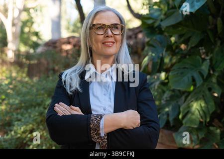 Lächelnde Geschäftsfrau mit überkreuzten Armen posiert in einem Park Stockfoto