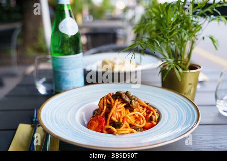 Ein Teller Spaghetti alla puttanesca, garniert mit grünen Oliven, Kapern und Kirschtomaten in einer reichhaltigen Tomatensoße. Das Gericht wird auf einem blauen Streifen serviert Stockfoto