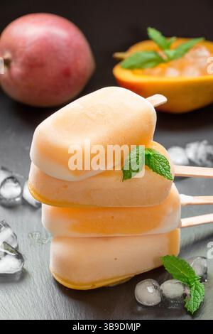 Spalte mit 4 Mangosorbet Eisbecher mischen Eis hinter Mango und geschnittener Mango in der Nähe von Eiswürfeln auf schwarzem Hintergrund. Mangosorbet Eisbecher mischen Eis Eis Eis Eis Eis Eis Eis Eis. Vertikal. Stockfoto
