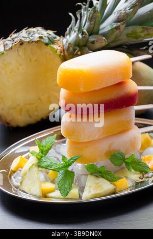 Spalte mit Sorbet-Eis-Eis-Eis von Ananasmangoherdbeere auf Silberplatte mit Eiswürfeln und Fruchtstücken auf schwarzem Hintergrund. Ananasmangoherdbeersorbet-Eis-Eis-Eis. Stockfoto