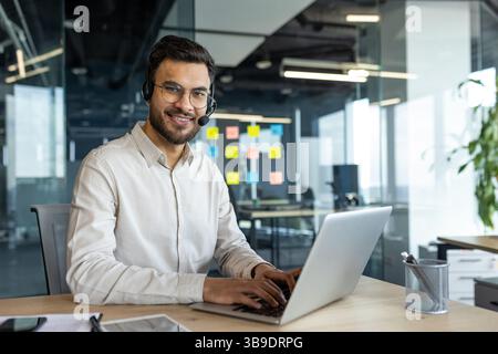 Porträt eines jungen erfolgreichen Firmenmitarbeiters mit Headset. Online-Kundendienstberater lächelnd und in die Kamera schaut, Laptop für Videoanrufe verwendet, im Büro arbeitet. Stockfoto