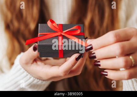 Elegante Geschenkbox Moment. Kleines schwarzes Gehäuse mit rotem Band. Die Hände der Frau lösen den Bogen. Feiertagsstimmung. Geben Sie das Konzept des Gebens vor. Stockfoto