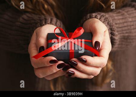 Frau, die ein kleines Geschenk hält. Black Box mit rotem Band. Geschenk zum Geburtstag oder Weihnachten. Urlaubsüberraschung in den Händen. Romantische festliche Geste. Stockfoto