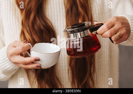 Frau, die Tee in eine Tasse gießt. Hände halten die Teekanne aus Glas. Heißes Kräutergetränk im Becher. Gemütlicher Wintertee. Warme, zwanglose Teeatmosphäre. Stockfoto