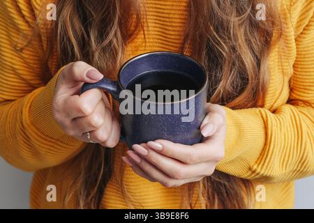 Frau hält Kaffeetasse. Heißes schwarzes Getränk in den Händen. Gelber Pullover und langes Haar. Warmer, gemütlicher Kaffee. Entspannter Vormittag zu Hause im Lifestyle-Hintergrund Stockfoto