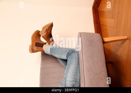 Beine einer Frau in Jeans mit braunen Stiefeln an den Füßen, die auf einem grauen Sofa liegt. Stockfoto
