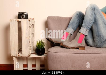 Die Beine einer Frau in Jeans mit grau-rosa Stiefeln auf einem grauen Sofa. Neben dem Sofa befinden sich Weißweinkisten mit einer Pflanze und einer Kamera als Dekoration Stockfoto