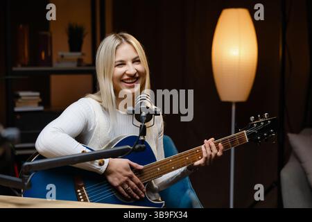 Lächelnde Musikerin singt und spielt Akustikgitarre, während sie einen Song in einem Heimstudio aufnimmt Stockfoto