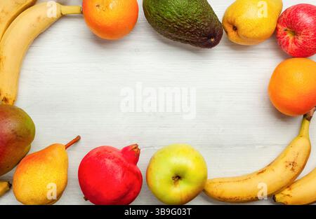 Buntes frisches Obst wie Bananen, Orangen, Avocado, Birnen, Äpfel, Mango und Granatapfel bilden einen Rahmen auf einem weißen Holztisch mit Kopierraum in der Mitte Stockfoto
