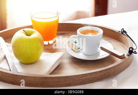 Holztablett mit frischem Frühstück mit Kaffee, Fruchtsaft und knusprigem Apfel, serviert in einem gemütlichen Hotelzimmer, das sich im warmen Sonnenlicht sonnt und für ein perfektes Erlebnis am Morgen sorgt Stockfoto