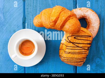Typisch italienisches Frühstück mit Espresso, Croissant, Donut und Saccottino, das auf einem blauen Holztisch serviert wird, bietet einen süßen und belebenden Start in den Tag Stockfoto