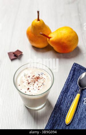 Joghurt mit geriebener Schokolade im Glas, zwei Birnen und Schokoladenchips, Löffel und blaue Serviette auf weißem Holztisch, gesundes Frühstück oder Snack Stockfoto