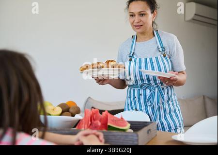 Eine Mutter mit Schürze serviert ihren Kindern beim Frühstück in einer gemütlichen Küche frisch gebackene Croissants. Der Tisch ist mit einer Vielzahl von Früchten gedeckt Stockfoto