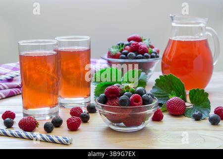 Links zwei Gläser Kompott Himbeeren, Erdbeeren, Heidelbeeren, in der Nähe Schüssel mit Beeren, rechts Karaffe Kompott auf einem hellen hölzernen Hintergrund verstreute Beeren. Kompott aus frischen Beeren. Horizontal. Stockfoto