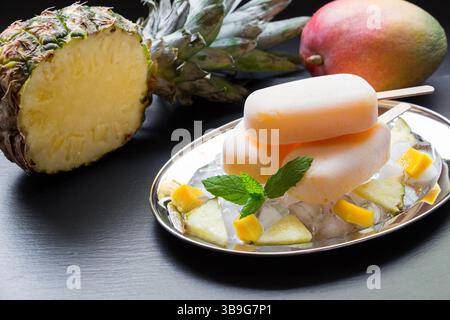 Exotisches Sorbeteis auf Silberplatte mit Eiswürfeln gehackte Ananasmangobiteile, Minze nahe der Hälfte der Ananas, Mango auf schwarzem Hintergrund. Ananas-Mangosorbet-Eis-Eis. Stockfoto