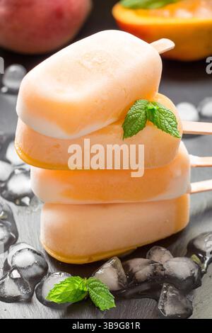 Drei Mangoheis-Eis-Eis stapeln sich auf einer dunklen Oberfläche übereinander. Sie sind von Eiswürfeln umgeben, was ein erfrischendes Erlebnis für einen warmen Sommertag ist. Stockfoto