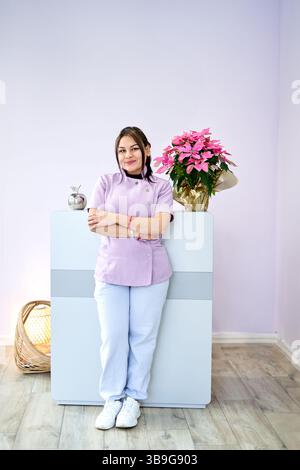 Ein professioneller Stylist steht selbstbewusst in einem gut beleuchteten Schönheitssalon. Der Raum ist mit hellvioletten Wänden und einer lebendigen Pflanze geschmückt, die einen TRA schafft Stockfoto