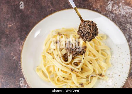 Tagliatelle-Pasta mit gehacktem schwarzen Trüffel auf dem Löffel, selektiver Fokus. Frisches italienisches Herbstrezept mit schwarzem Trüffel auf braunem Tisch in eleganter r Stockfoto
