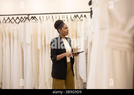 Eine Frau steht in einer Brautboutique und stöbert durch eine Kollektion eleganter weißer Brautkleider. Sie hält ein Tablet in der Hand und konzentriert sich auf die Auswahl der perfekten Dres Stockfoto