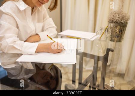 Eine Frau in einem weißen Hemd schreibt auf Papier an einem Glastisch. Reflektionen auf der Oberfläche verleihen der Oberfläche Tiefe, mit minimalistischem Dekor, einschließlich einer Pflanze und einem Bleistift Stockfoto