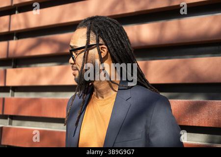 Ein kubanischer Geschäftsmann mit langen Dreadlocks, Sonnenbrille und Blazer, sonnt sich im Sonnenlicht. Das Umgebungslicht und die Holzkulisse sorgen für ein warmes A Stockfoto
