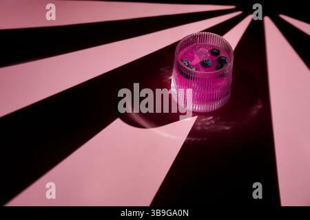 In einem gerippten Glas sitzt ein leuchtend rosafarbenes Getränk, das mit Heidelbeeren garniert ist und dramatische Schatten auf einer rosafarbenen Oberfläche wirft. Der künstlerische Kontrast sorgt für visuelle Effekte Stockfoto
