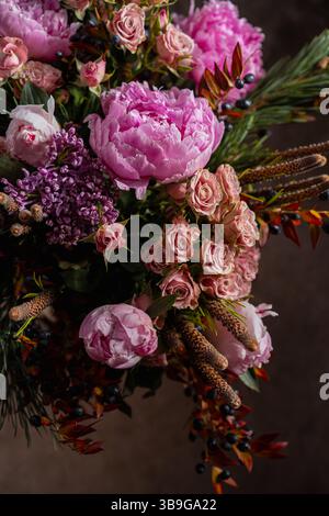 Ein luxuriöses Blumenarrangement mit zarten rosa Pfingstrosen, zarten Fliederblüten und rosa Rosen mit dunklen Beeren und Kiefernlaub Stockfoto