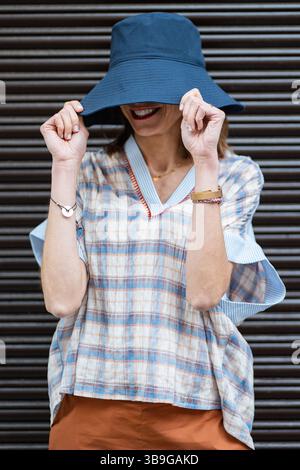 Eine Modefrau versteckt ihr Gesicht spielerisch mit einem blauen Hut mit breiter Krempe, trägt ein kariertes Hemd und Armbänder. Dieses legere Sommeroutfit ist perfekt für Stockfoto