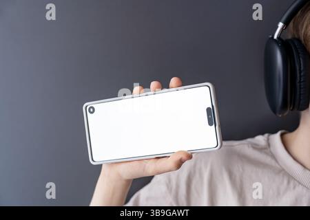 Ein Teenager hält ein Smartphone mit einem leeren weißen Bildschirm im Querformat, während er Kopfhörer trägt. Der klare, minimalistische Hintergrund ist perfekt für Mockups Stockfoto