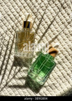 Zwei elegante Tropfer-Kosmetikflaschen mit goldenen und grünen Seren liegen auf strukturiertem weißem Stoff im Badezimmer. Natürliches Licht wirft künstlerische Schatten, cr Stockfoto
