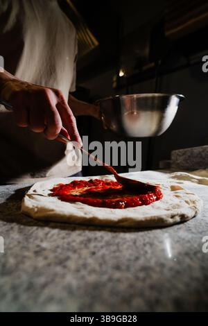 Eine Person, die frische Tomatensauce auf einem Pizzateig in einer schwach beleuchteten Küche verteilt. Der Fokus liegt auf dem Prozess der Herstellung einer köstlichen hausgemachten Pizza aus Stockfoto
