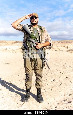 Ein Soldat steht in einer Wüstenumgebung, trägt Tarnkleidung und Sonnenbrille. Er hält ein Gewehr und grüßt, zeigt Respekt und Professionalität Stockfoto