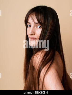 Ein Porträt einer jungen Frau mit langen braunen Haaren, die in einem Atelier auf beigefarbenem Hintergrund die Kamera über ihre Schulter schaut. Ihr Blick ist subtil und fügt de hinzu Stockfoto