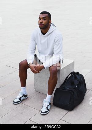 Ein junger männlicher Sportler sitzt draußen auf einem Betonblock. Er trägt einen weißen Hoodie und Turnschuhe, neben ihm eine Sporttasche. Entspannt, entspannt Stockfoto