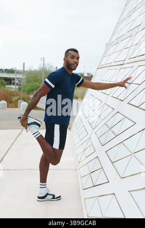Ein schwarzer Sportler streckt sein Bein, während er sich gegen eine geometrische Wand lehnt. In Sportkleidung zeigt er Outdoor-Fitness-Engagement Stockfoto