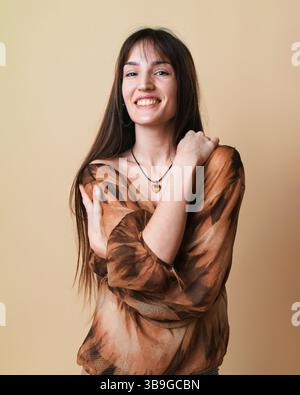 Porträt einer jungen Frau auf beigefarbenem Hintergrund, aufgenommen in einem Studio. Sie lächelt und trägt eine braune gemusterte Bluse, die ein Jo ausdrückt Stockfoto