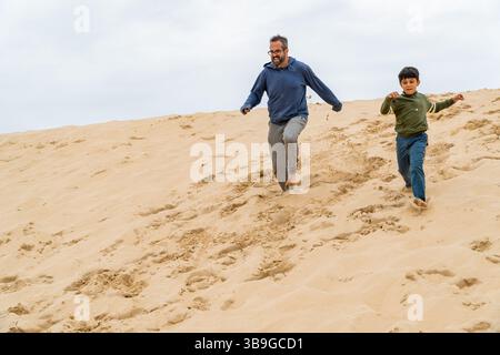 Ein fröhlicher Vater und Sohn laufen barfuß die Düne von Pilat an einem bewölkten Tag hinunter und teilen einen Moment der Bindung, des Spaßes und des Lachens, um das Wesen festzuhalten Stockfoto